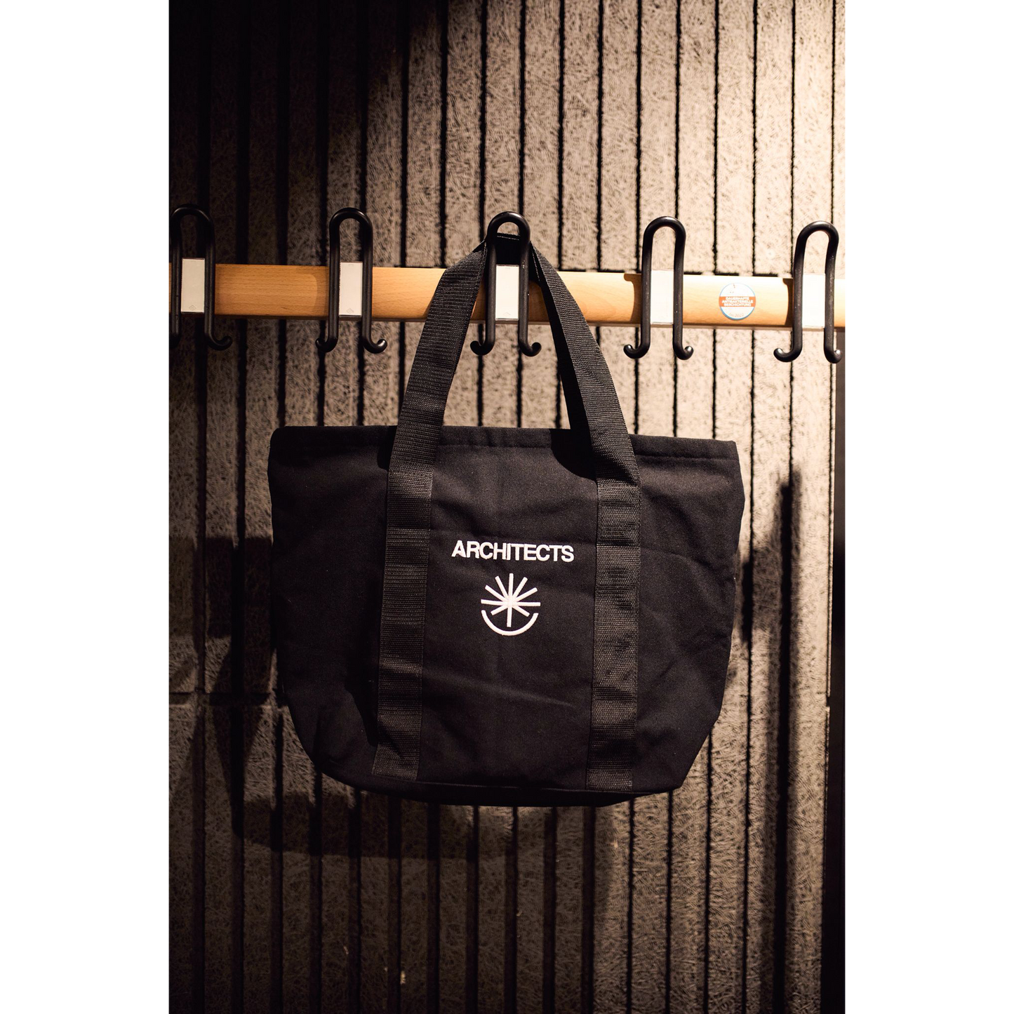 Black bag with 'ARCHITECTS' logo hanging on a wooden rack against a textured wall.