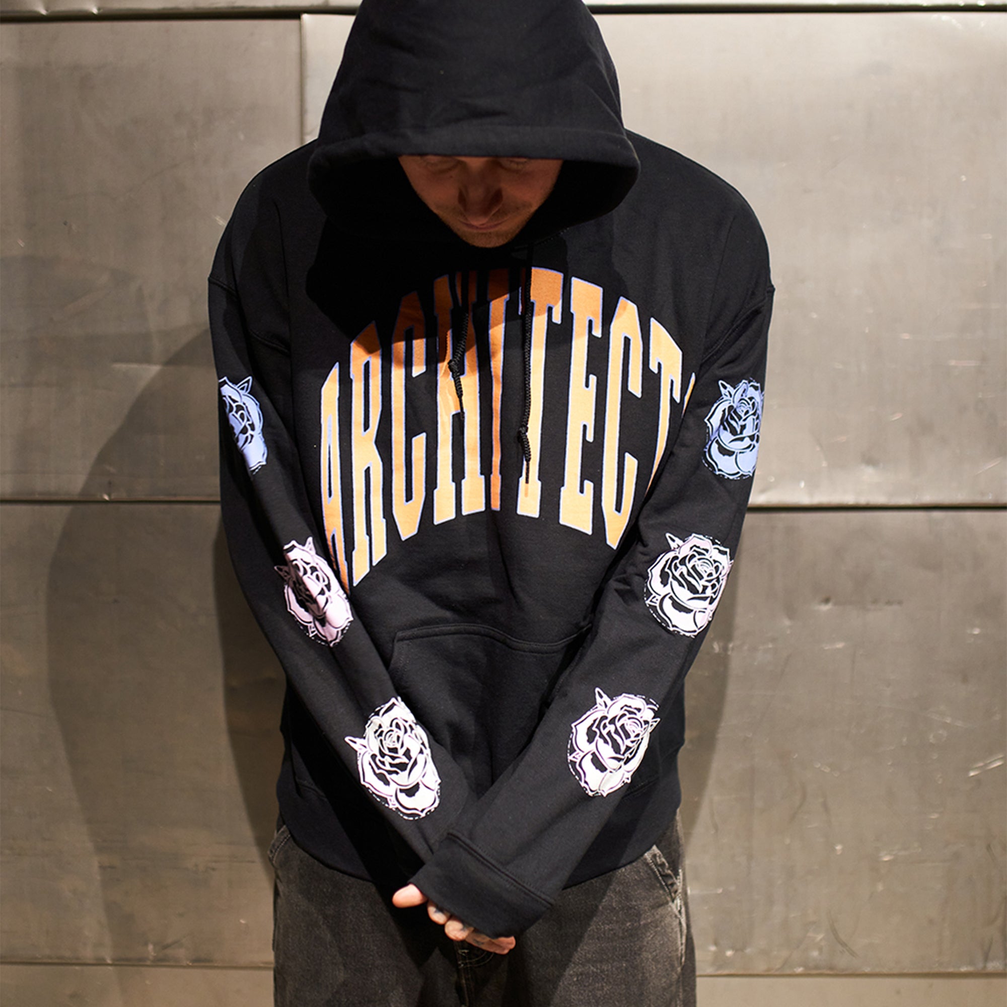 Black regular-fit unisex hoodie featuring an orange Architects print across the front, with blue, pink, and white rose prints along the sleeves.