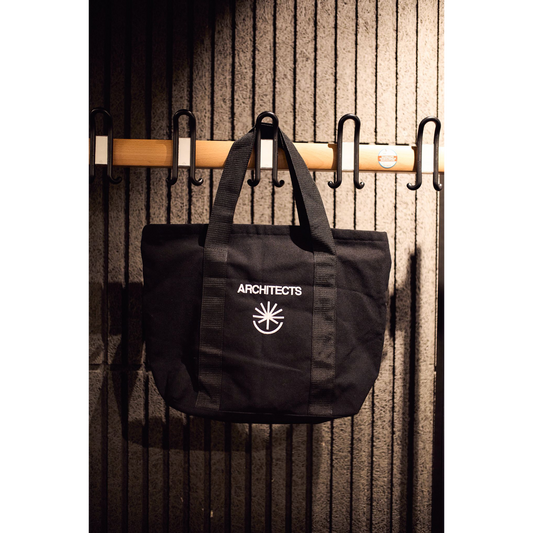 Black bag with 'ARCHITECTS' logo hanging on a wooden rack against a textured wall.